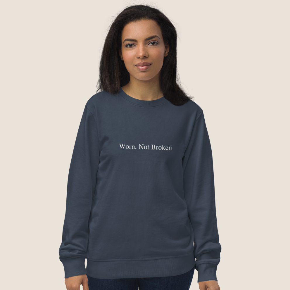 A woman modeling a slate blue crewneck sweatshirt with the white printed text 'Worn, Not Broken' on the chest, set against a solid light beige background.