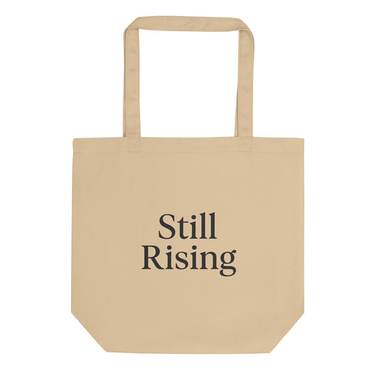 Beige tote bag with 'Still Rising' text on a white background